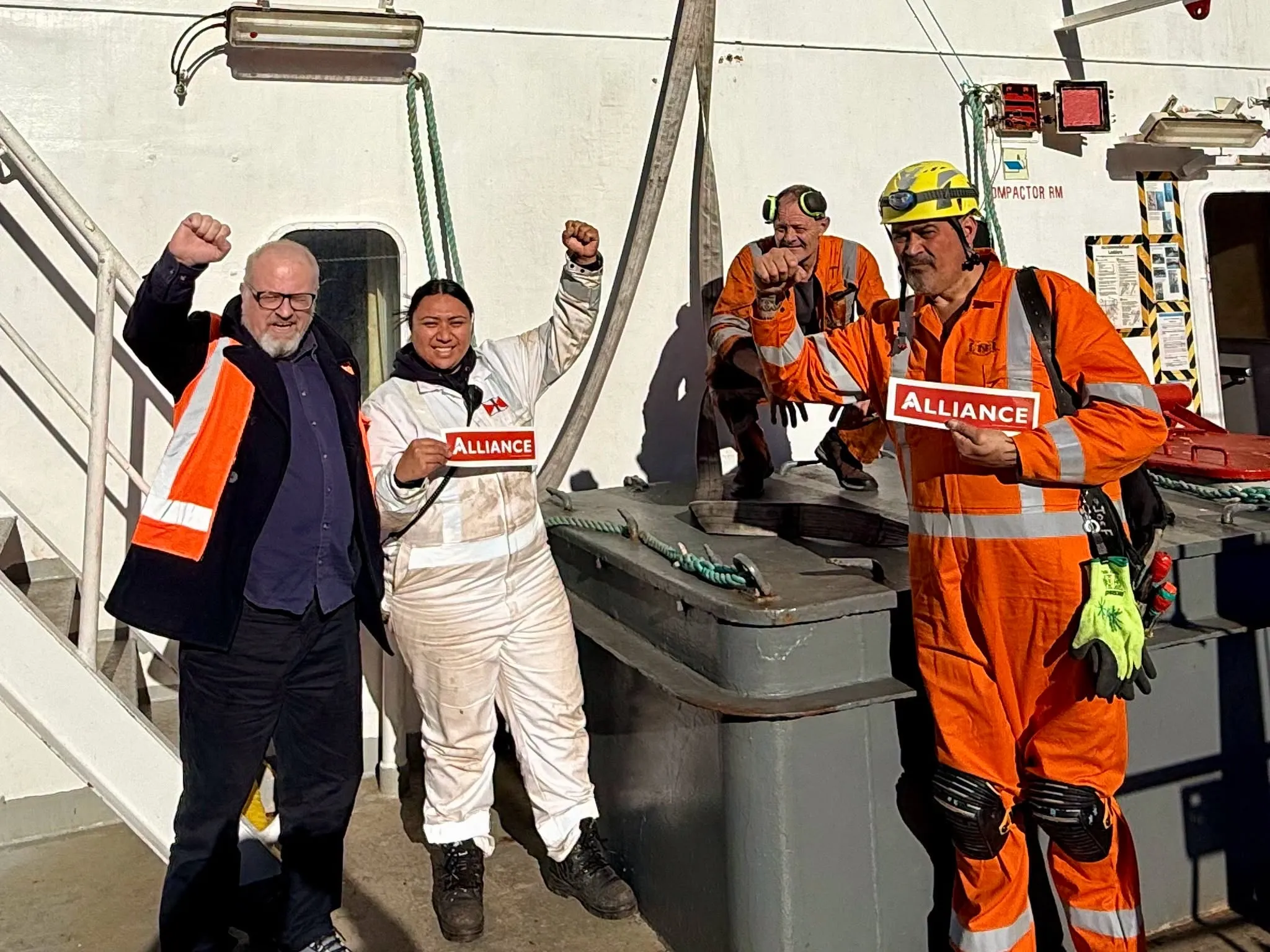 Maritime union crew members on a ship’s deck raise their fists and hold red Alliance signs