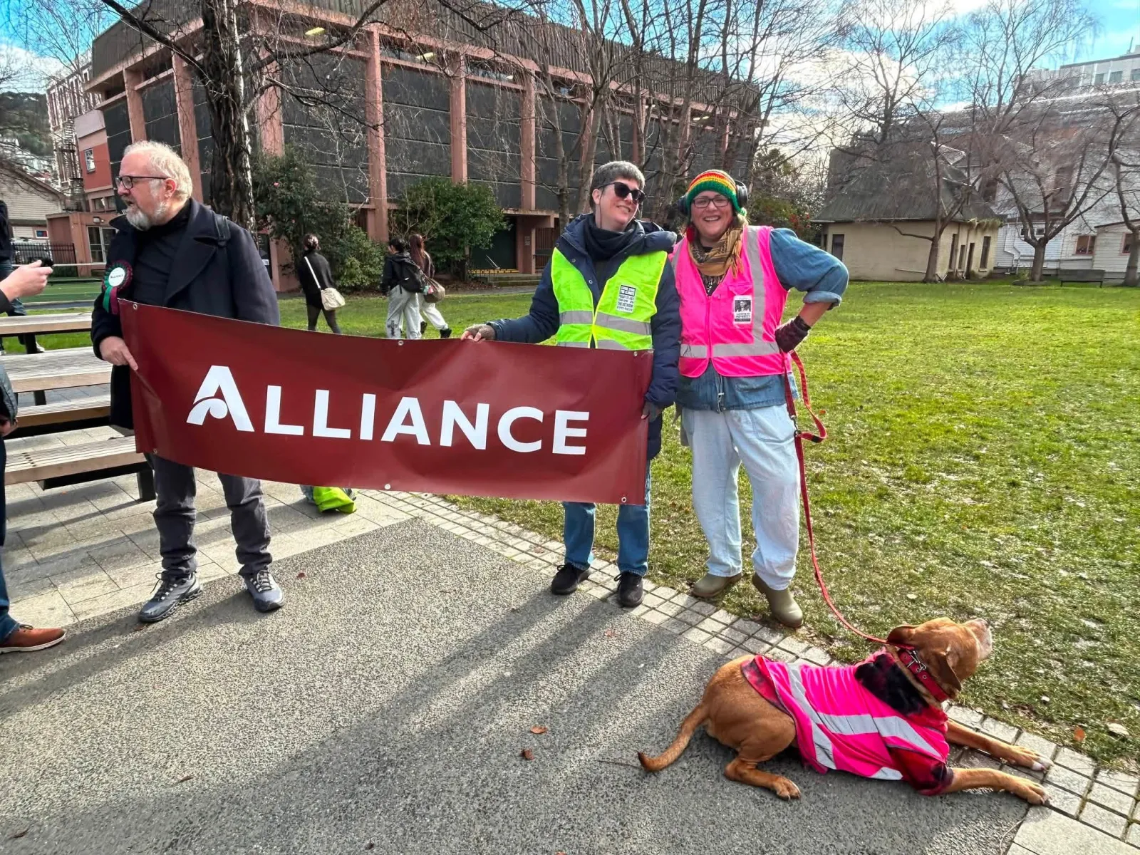 Victor Billot and Anna Knight with an Alliance banner outdoors, with a dog wearing a pink high-visibility vest beside them