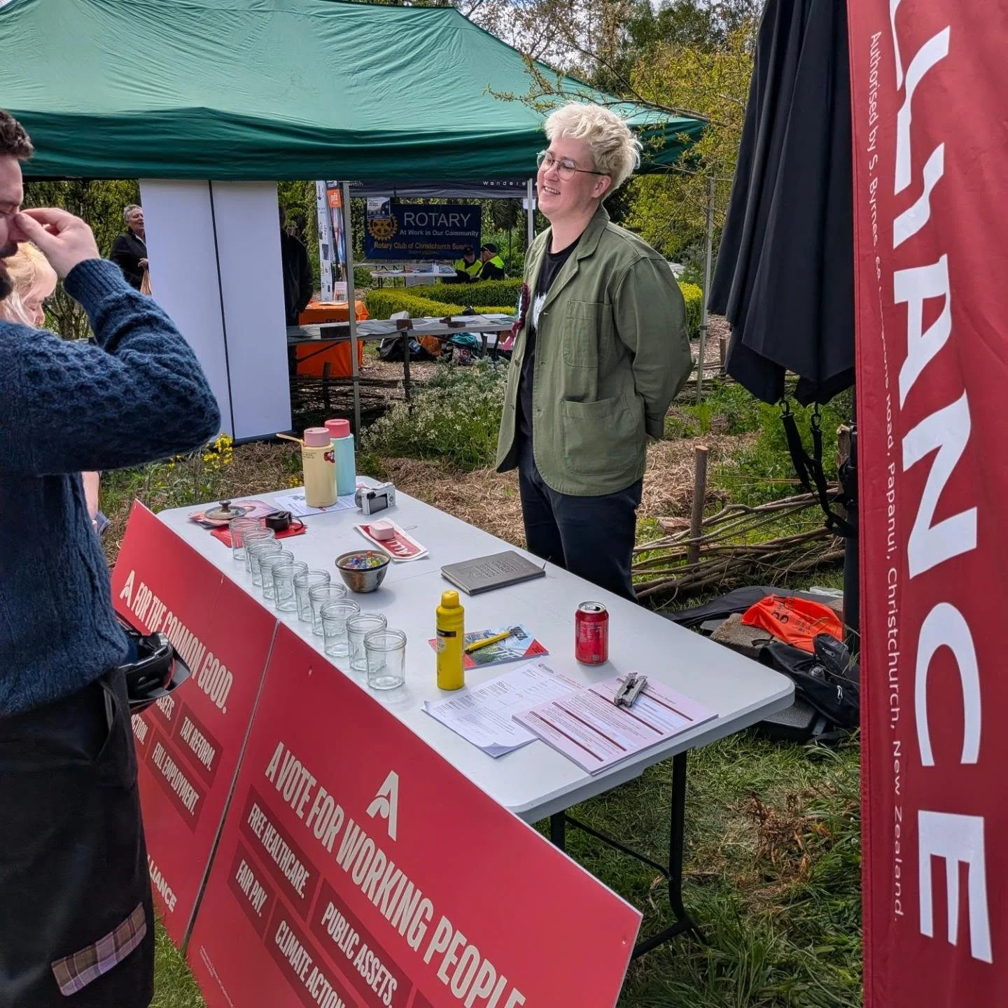 Kyle Turnbull talking with a visitor at an outdoor Alliance stall, beside campaign signs and leaflets