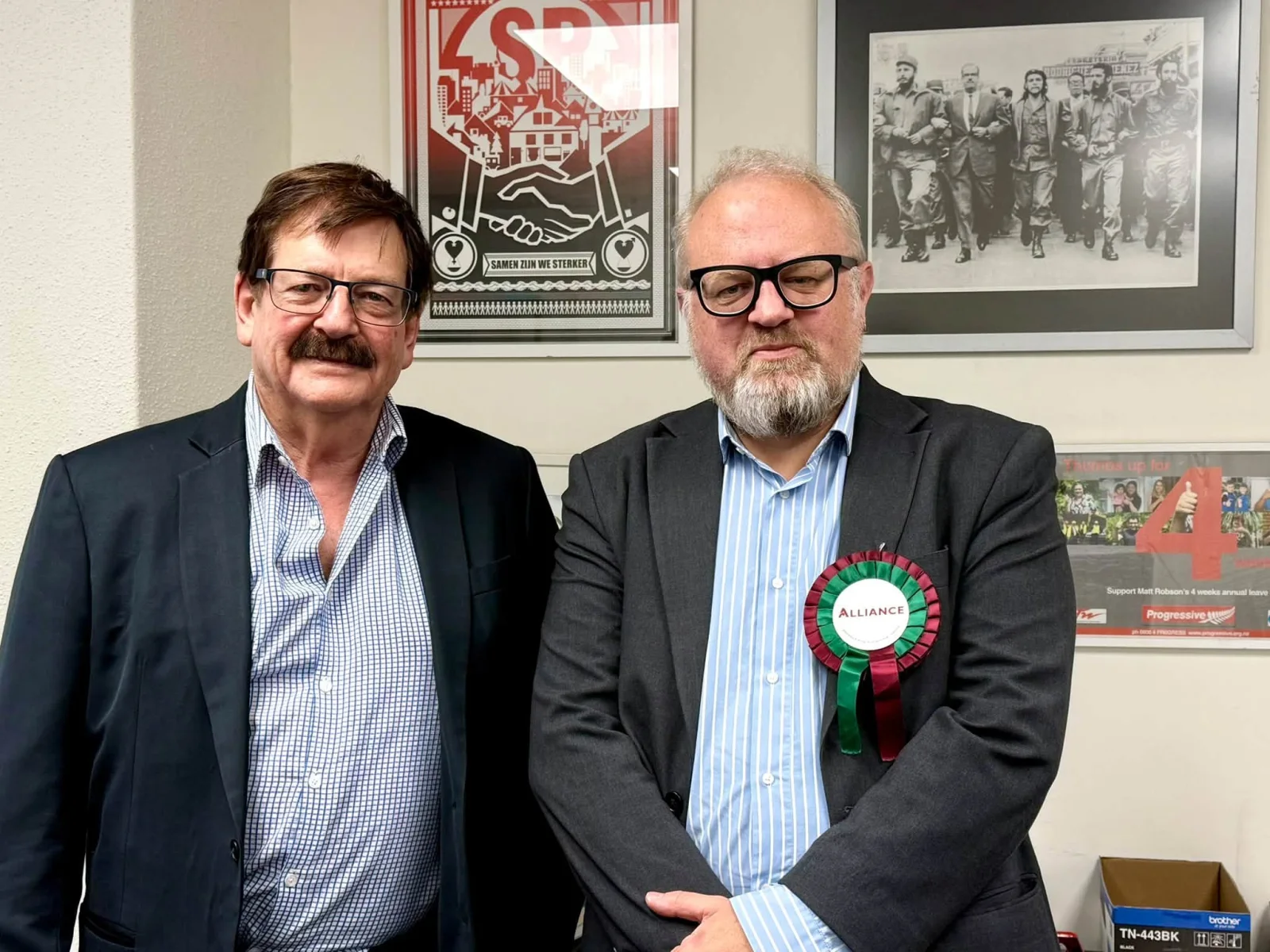 Matt Robson and Victor Billot standing together in front of framed political posters and a historical photograph