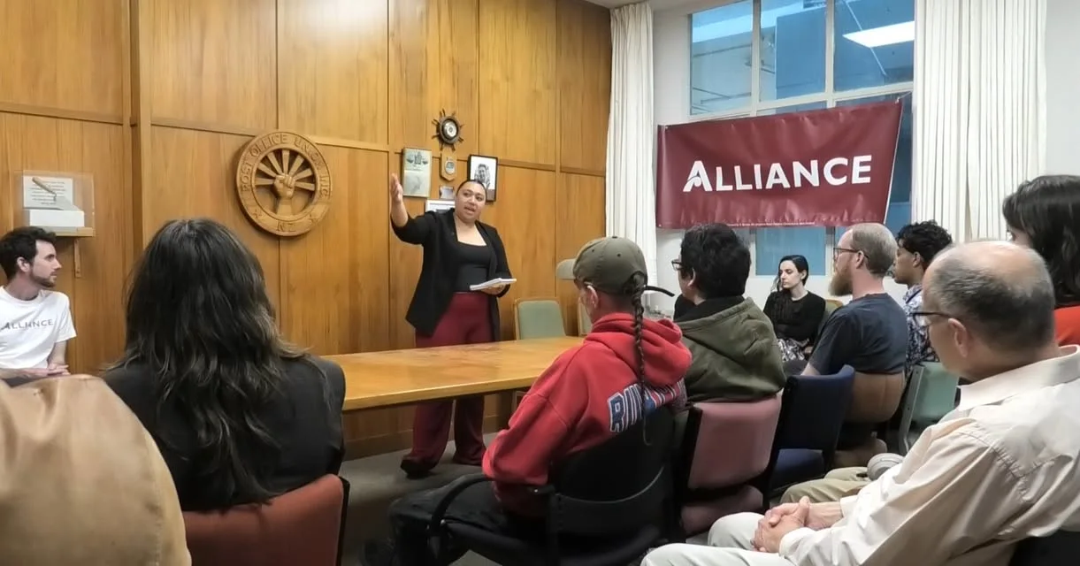 Victoria Ale speaking to a seated group at a meeting, with an Alliance banner behind her