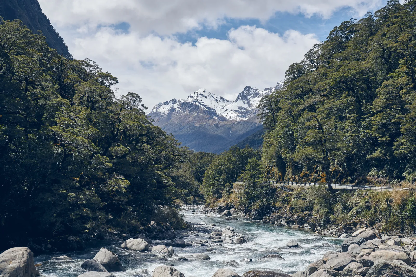 A river through native forest beneath snow-covered mountains on the road to Milford Sound.