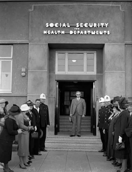 Michael Joseph Savage standing at the entrance to the Social Security and Health Departments building, with officials lining the steps.