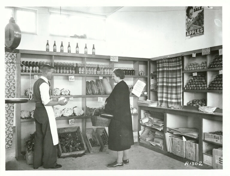 A greengrocer serves a customer beside shelves of fruit and vegetables in a historic New Zealand shop.