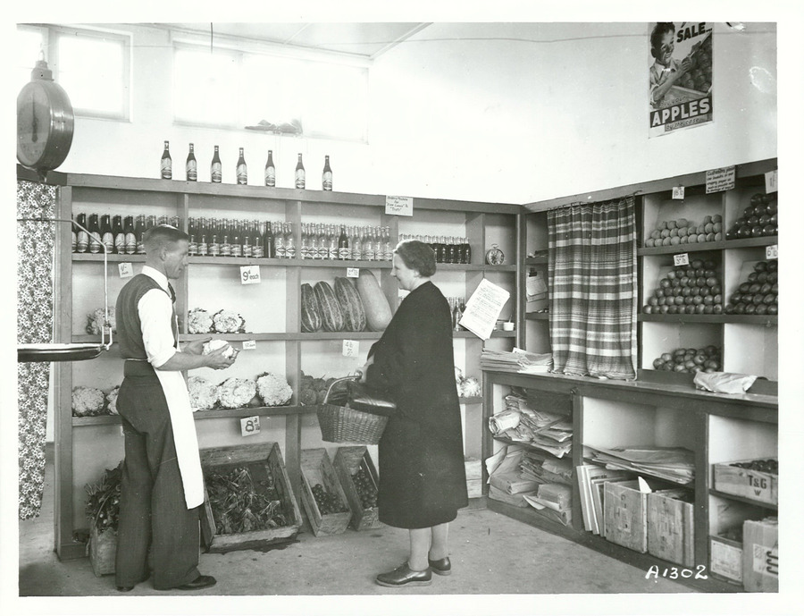 A greengrocer serves a customer beside shelves of fruit and vegetables in a historic New Zealand shop.