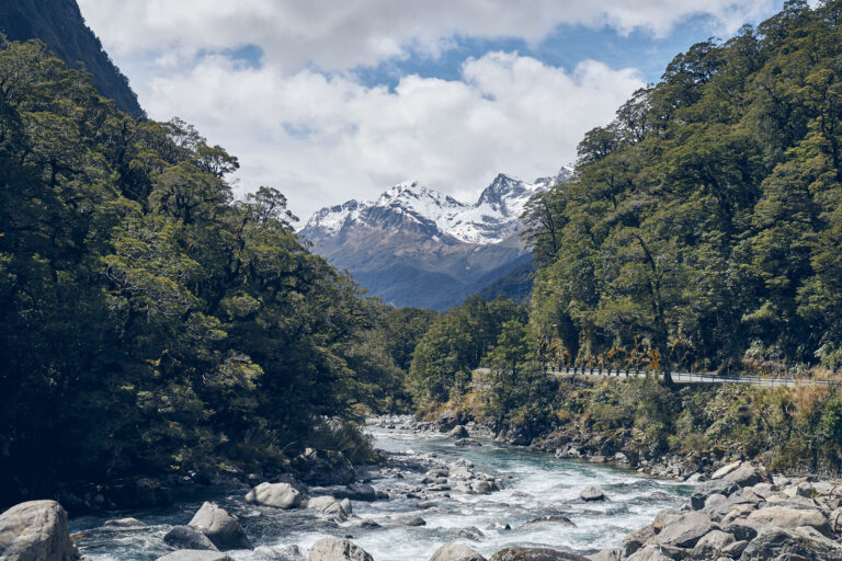 A river through native forest beneath snow-covered mountains on the road to Milford Sound.